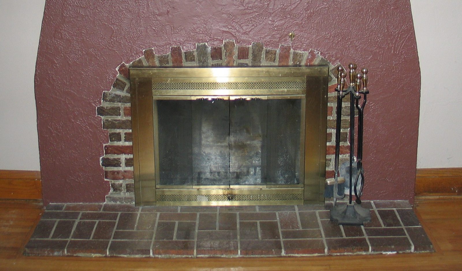 A home fireplace with brass-framed glass doors, a brick surround and hearth, and a set of fireplace tools standing beside it