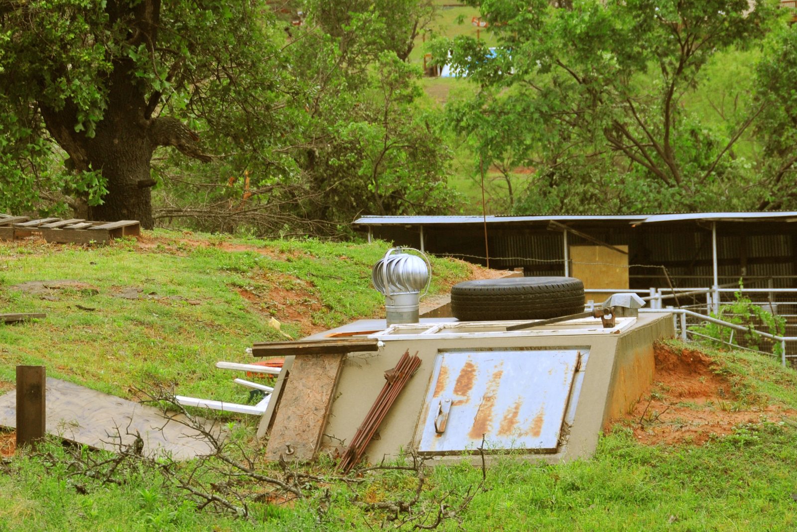 An above-ground tornado storm shelter standing beside a home in Oklahoma, with debris scattered around it