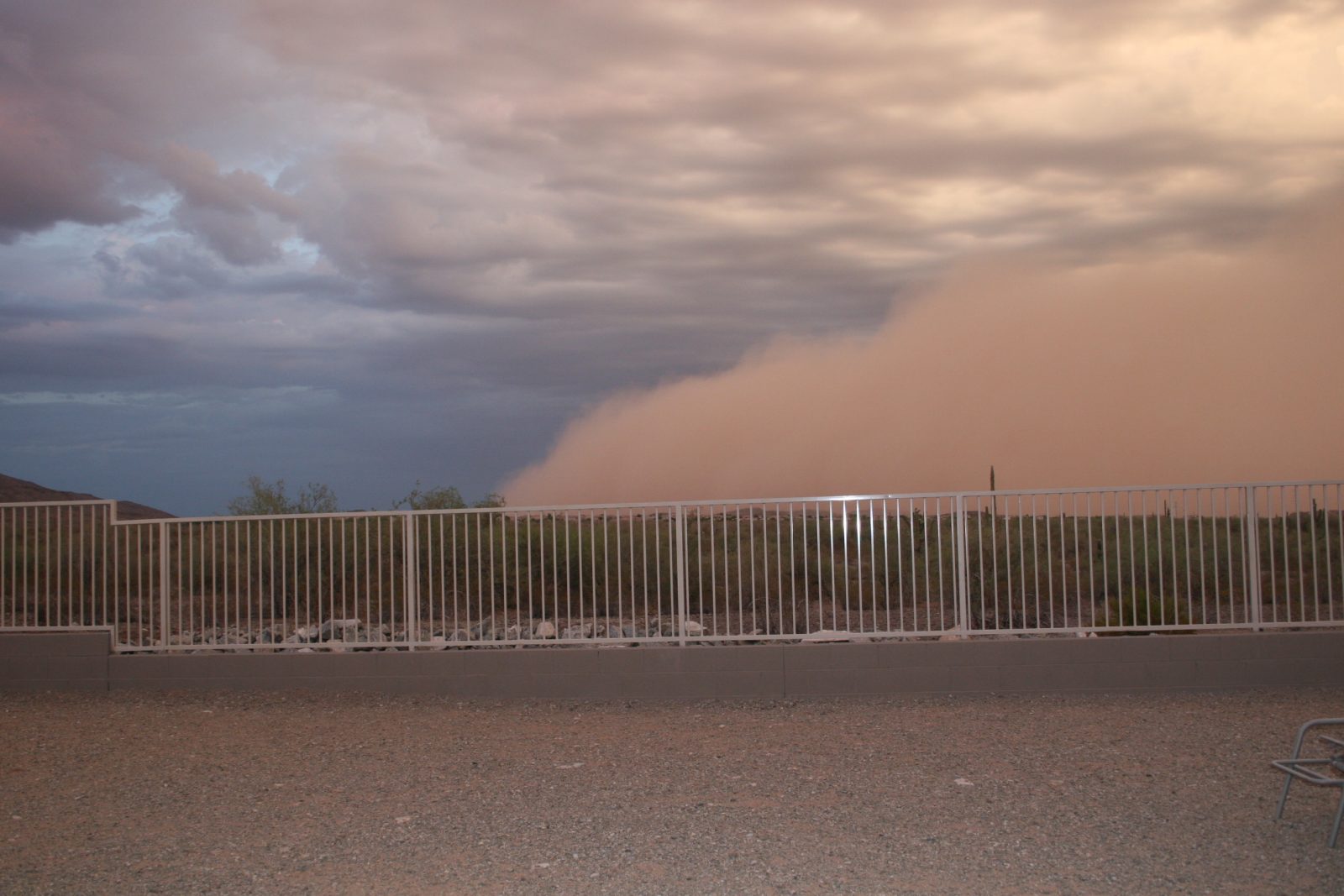 A towering wall of reddish-brown dust from a haboob advancing over a residential fence in Ahwatukee, Phoenix, Arizona, with dark storm clouds above it